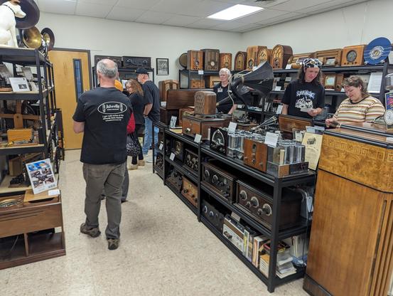 Right side of the museum with racks of radio equipment, mostly older table top and floor model receivers, some with vacuum tubes prominently displayed. There are also a few people in the picture.