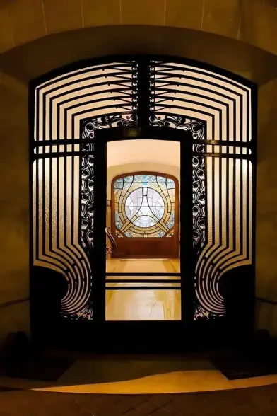 An Art Deco doorway in strong yet elegant forged iron, lit from a foyer behind. Perfectly framed in the door on the rear wall of the foyer is an Art Nouveau stained glass window showing a transition of eras